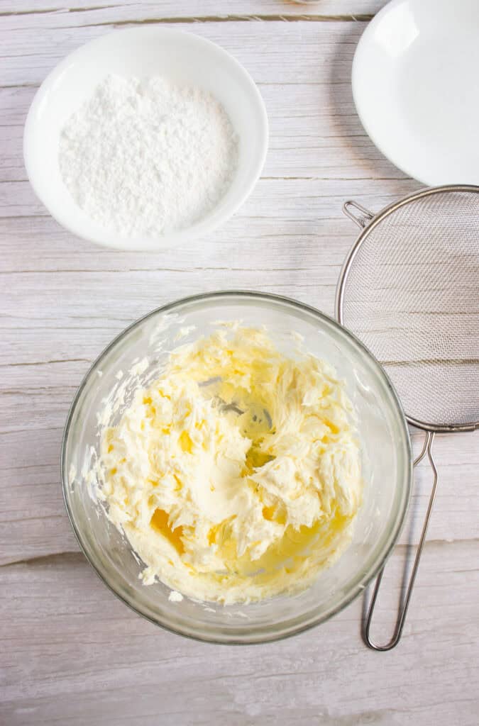 A bowl of whipped butter, a bowl of powdered sugar, a plate, and a sifter on a light surface—perfect for preparing Cream Cheese Frosting to top your favorite Pumpkin Cookies Recipe.