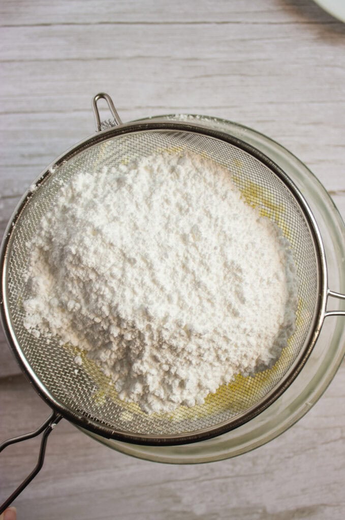 Flour being sifted through a metal sieve over a glass bowl on a light wooden surface, a perfect start for making soft pumpkin cookies or preparing the base for your favorite pumpkin cookies recipe with cream cheese frosting.