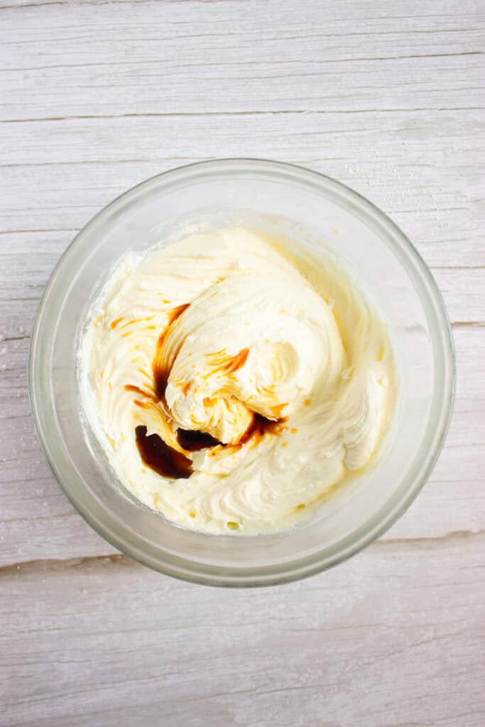 Glass bowl with whipped cream cheese and vanilla extract on a white wooden surface, perfect for making Cream Cheese Frosting to top your favorite Pumpkin Cookies.