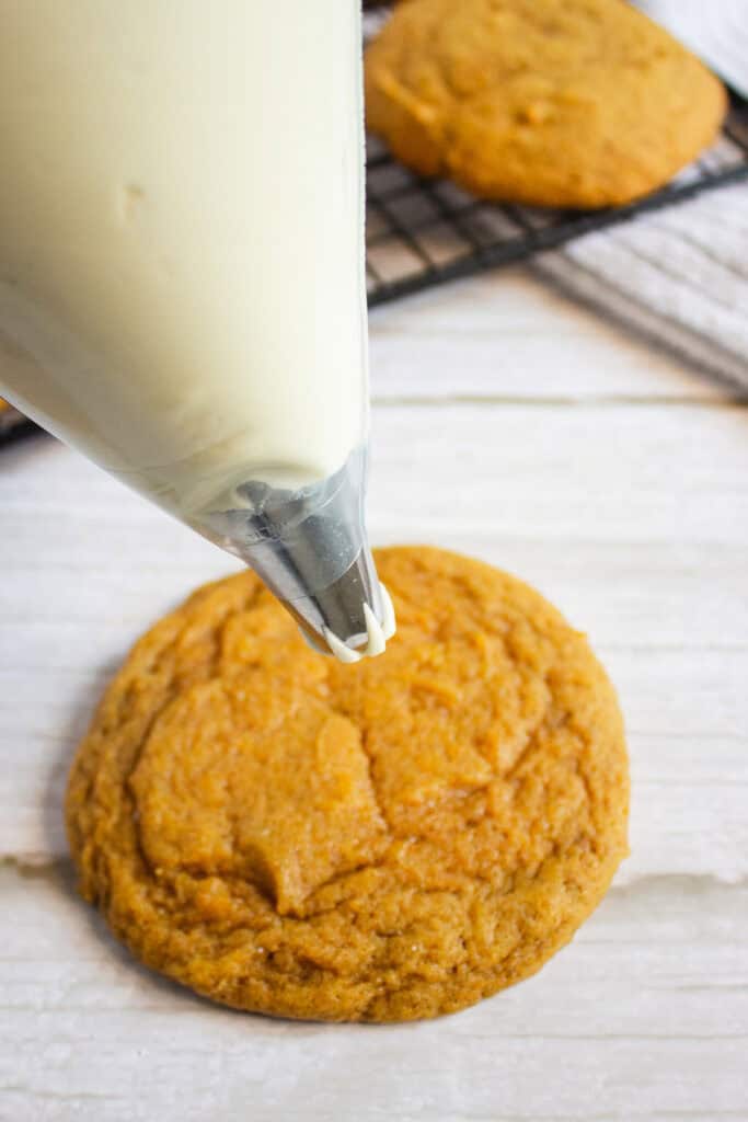 Piping creamy white icing onto a round pumpkin cookie, with more pumpkin cookies cooling on a rack in the background, makes for the perfect homemade pumpkin dessert.