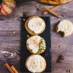 Three pumpkin cookies with cream cheese frosting rest on a slate board, surrounded by cinnamon sticks, star anise, and pumpkin seeds.