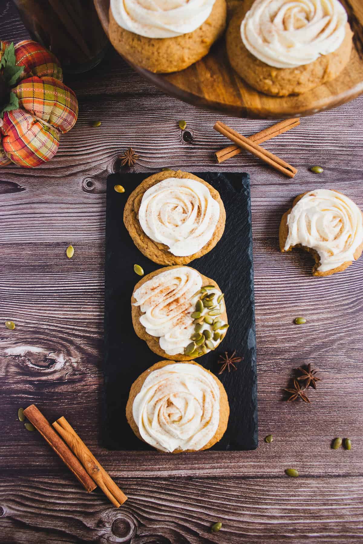 Three pumpkin cookies with cream cheese frosting rest on a slate board, surrounded by cinnamon sticks, star anise, and pumpkin seeds.