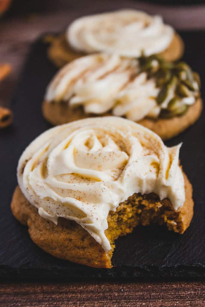 A delicious pumpkin cookie with creamy white cream cheese frosting and a bite taken out, displayed in front of two more frosted pumpkin cookies.
