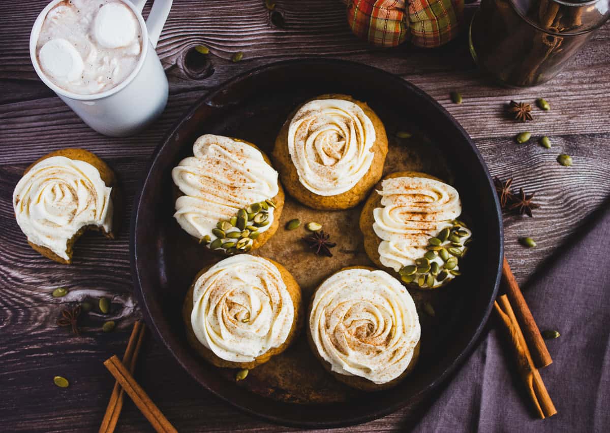 Five frosted pumpkin cupcakes on a round tray, with spices, pumpkin seeds, and a mug of hot chocolate nearby&mdash;just like your favorite Pumpkin Cookies Recipe topped with luscious cream cheese frosting.
