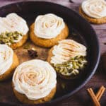 Round pumpkin cookies with swirled cream cheese frosting, some topped with pumpkin seeds, are arranged on a rustic tray alongside aromatic cinnamon sticks-perfect for trying a cozy new pumpkin cookies recipe.