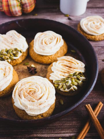 Round pumpkin cookies with swirled cream cheese frosting, some topped with pumpkin seeds, are arranged on a rustic tray alongside aromatic cinnamon sticks-perfect for trying a cozy new pumpkin cookies recipe.