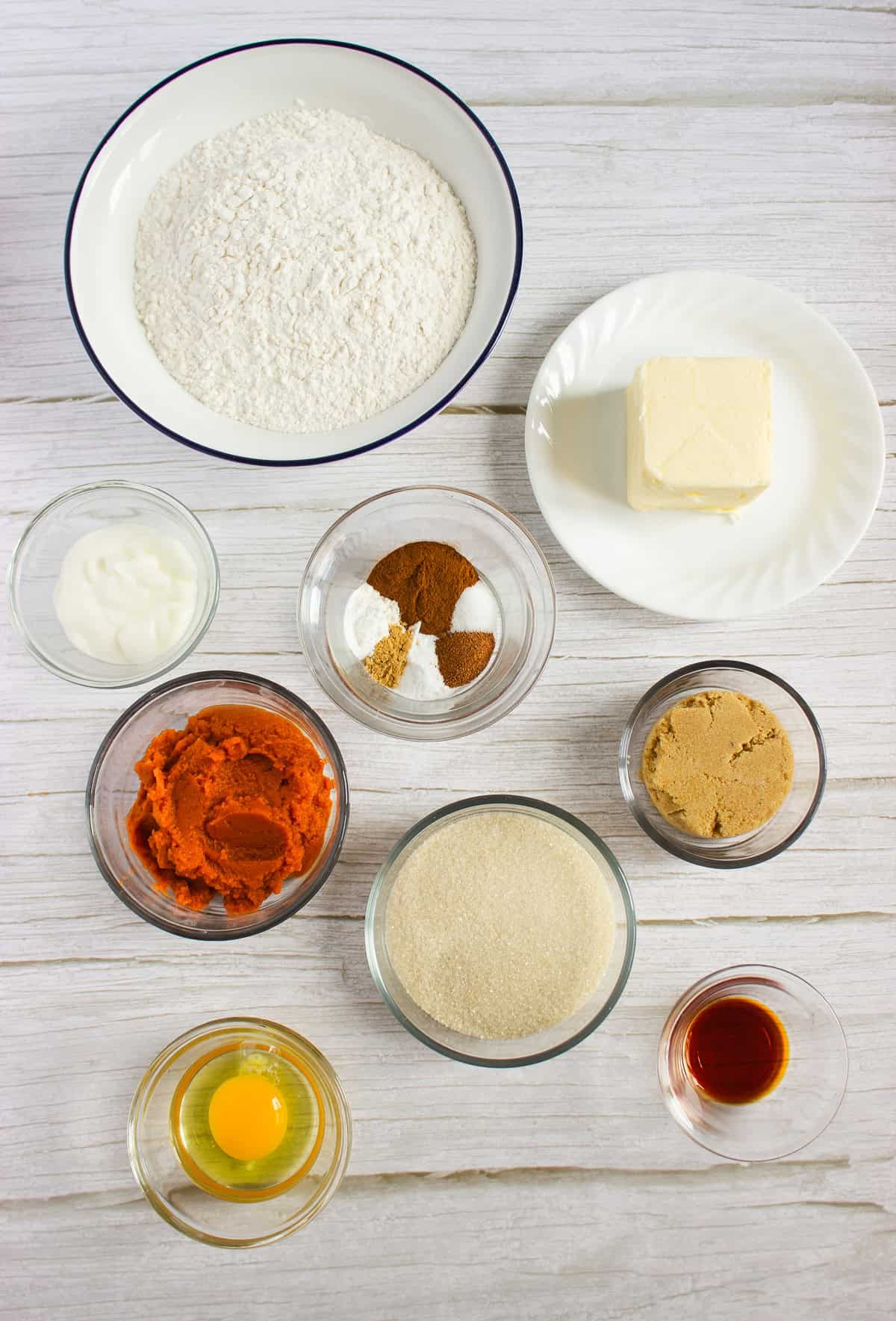 Bowls of flour, butter, sugar, brown sugar, pumpkin puree, spices, egg, vanilla, and yogurt on a white table—everything you need to start a delicious pumpkin cookies recipe or whip up soft Pumpkin cookies with irresistible cream cheese frosting.