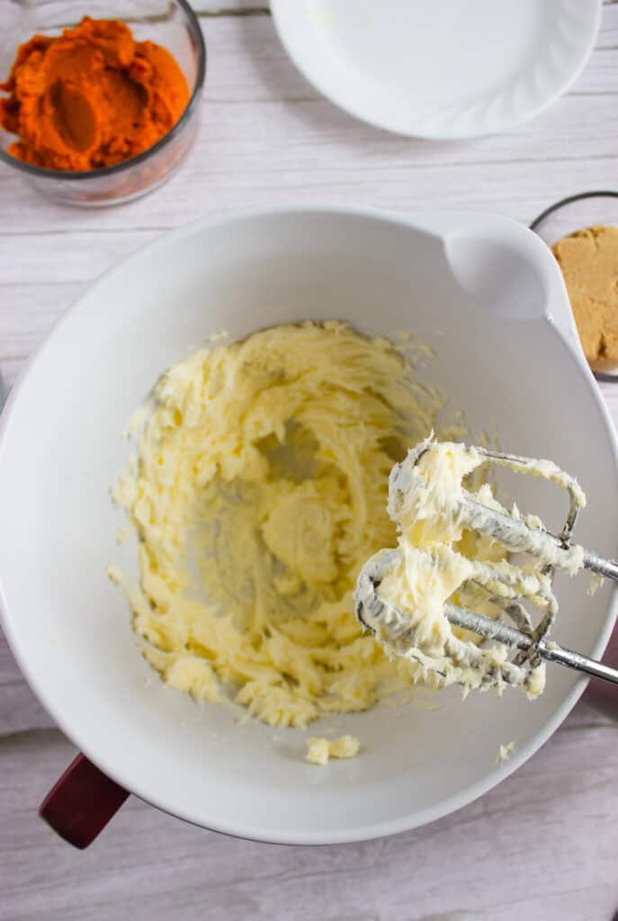 Creamed butter and sugar in a bowl with a hand mixer, ready to be combined with pumpkin puree and brown sugar nearby for delicious pumpkin cookies. Perfect finished with a dollop of cream cheese frosting.