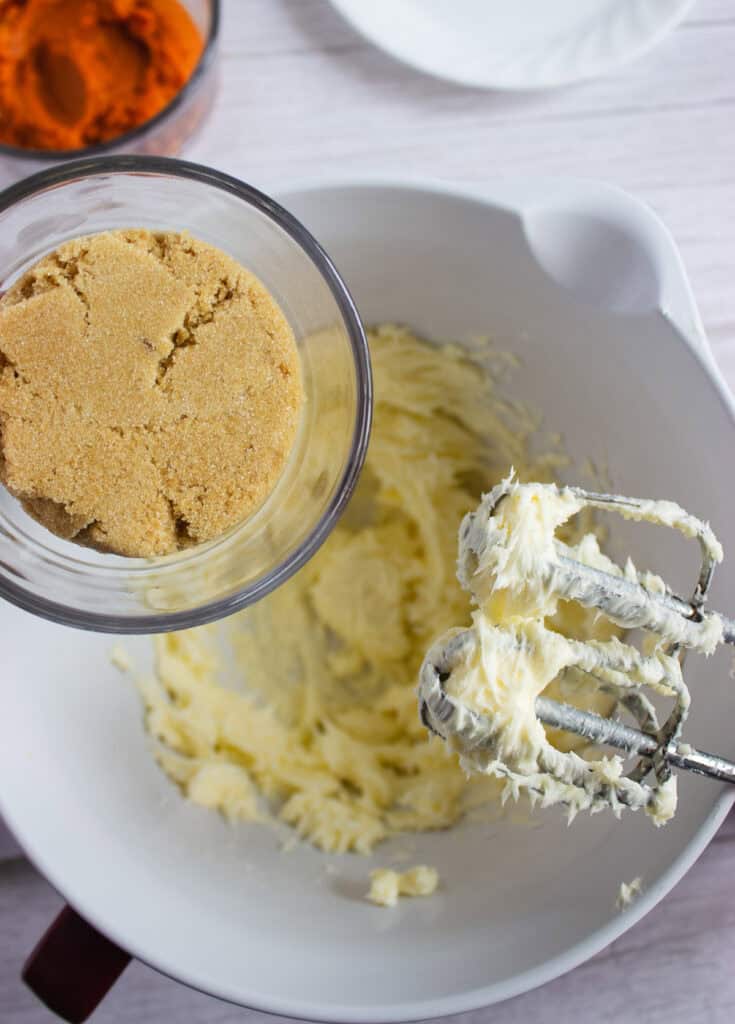 A glass of brown sugar held above a bowl of creamed butter with an electric mixer on the side, perfect for preparing the base of a delicious Cream Cheese Frosting for your favorite Pumpkin Cookies Recipe.