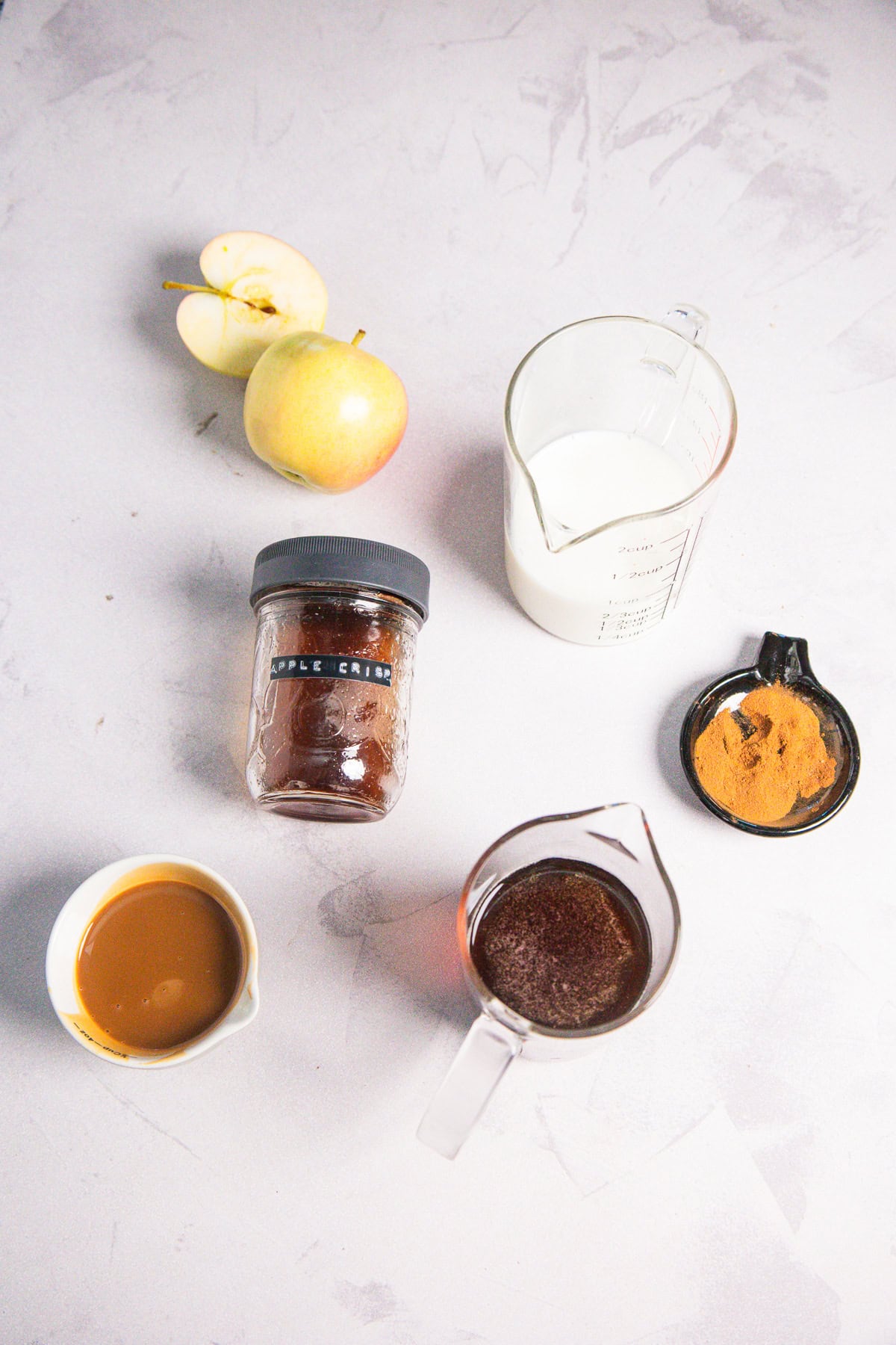 Sliced apple, whole apple, milk, cinnamon, apple crisp jar, caramel, and syrup on a light surfaceโeverything you need for the perfect Caramel Apple Iced Coffee moment.