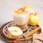 A Caramel Apple Iced Coffee latte topped with foam, caramel drizzle, apple slices, and a cinnamon stick on a wooden tray.