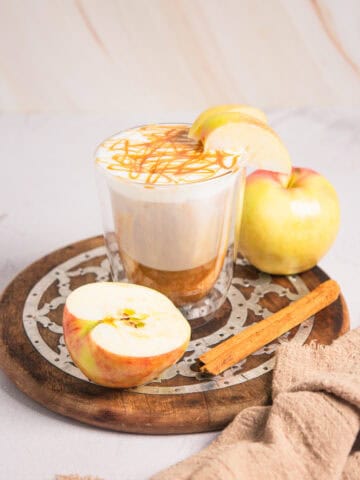 A Caramel Apple Iced Coffee latte topped with foam, caramel drizzle, apple slices, and a cinnamon stick on a wooden tray.