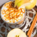 A caramel-drizzled latte topped with apple slices, surrounded by apples and cinnamon sticks on a tray, brings all the cozy flavors of a classic caramel apple. This inviting drink is perfect for fans of Caramel Apple Iced Coffee or anyone craving a sweet autumn treat.