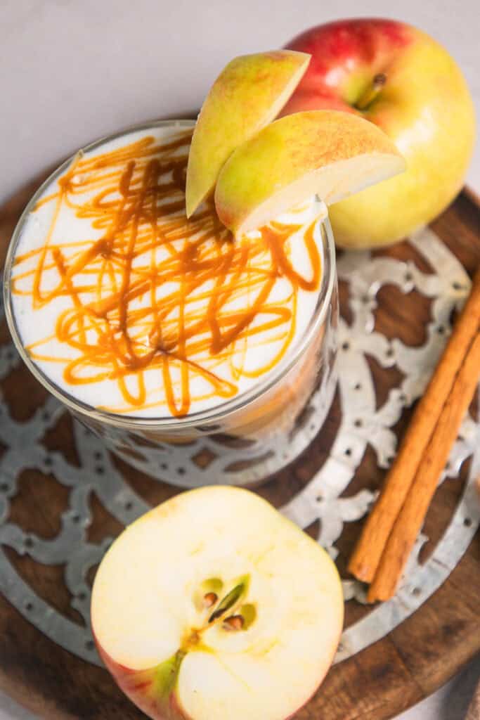 A caramel-drizzled latte topped with apple slices, surrounded by apples and cinnamon sticks on a tray, brings all the cozy flavors of a classic caramel apple. This inviting drink is perfect for fans of Caramel Apple Iced Coffee or anyone craving a sweet autumn treat.