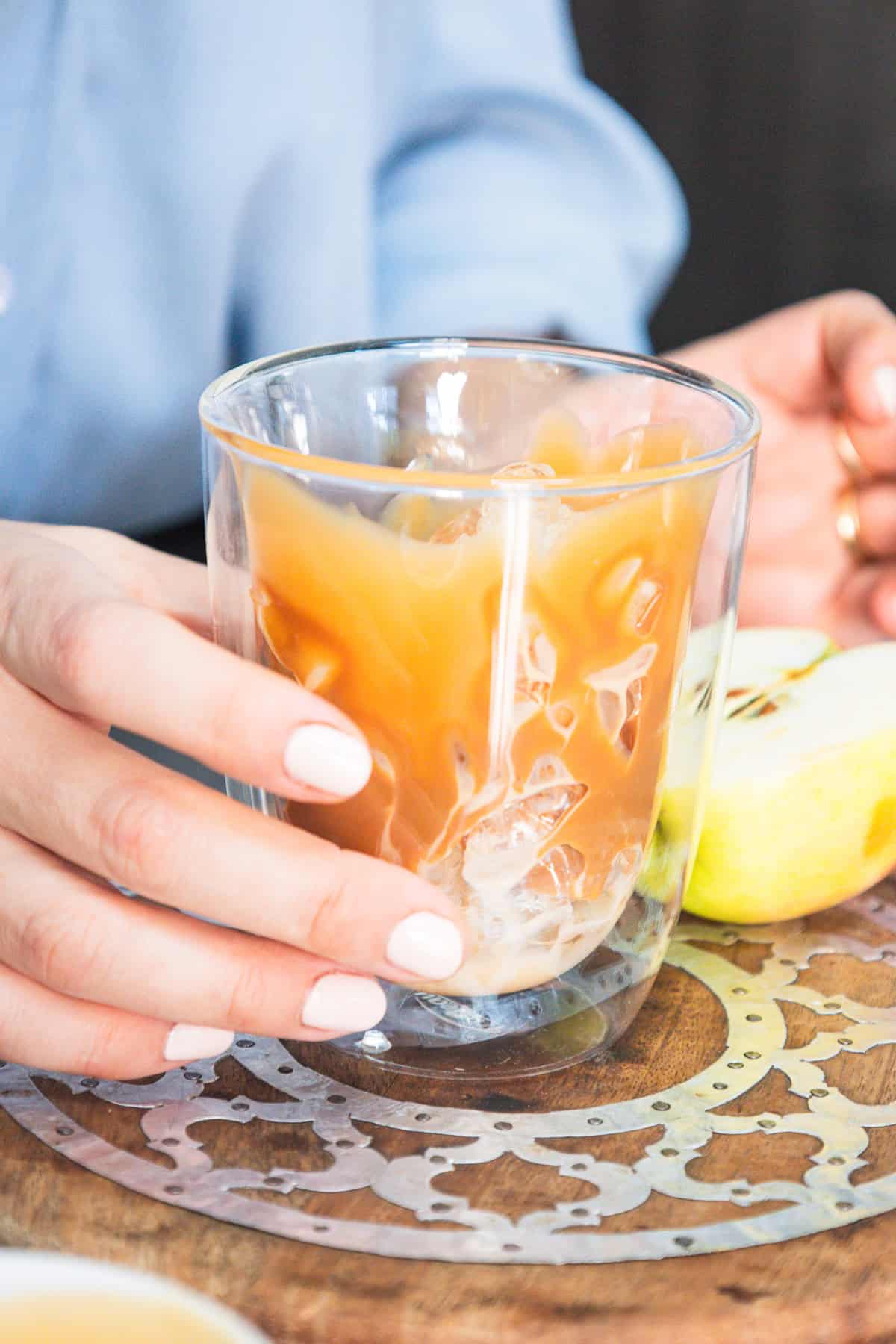 Hand holding a glass of Caramel Apple Iced Coffee next to a sliced green apple on a wooden table.