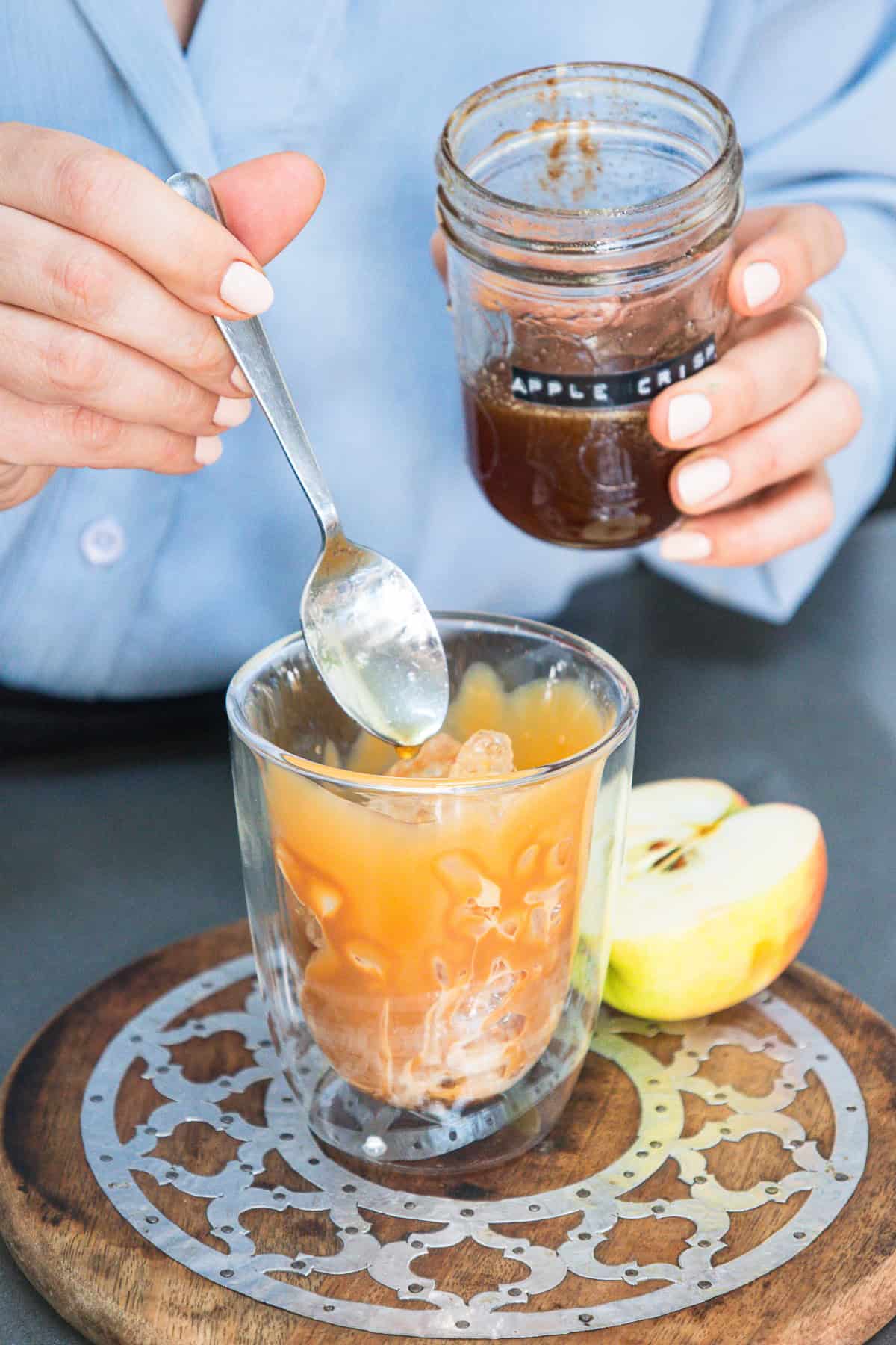 A person adds syrup from a jar to an iced apple drink in a glass, creating a tempting Caramel Apple Iced Coffee next to sliced apples on a wooden board.