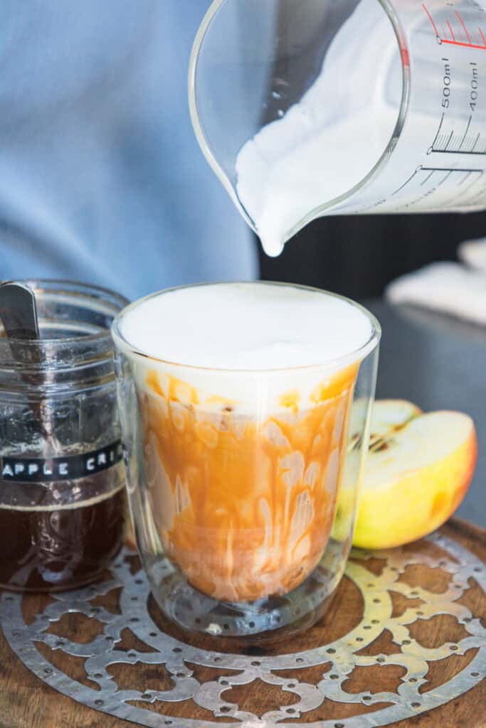 Milk is being poured into a glass of tea, with apple slices and a jar labeled โapple crispโ nearby, evoking the cozy flavors of Caramel Apple Iced Coffee.