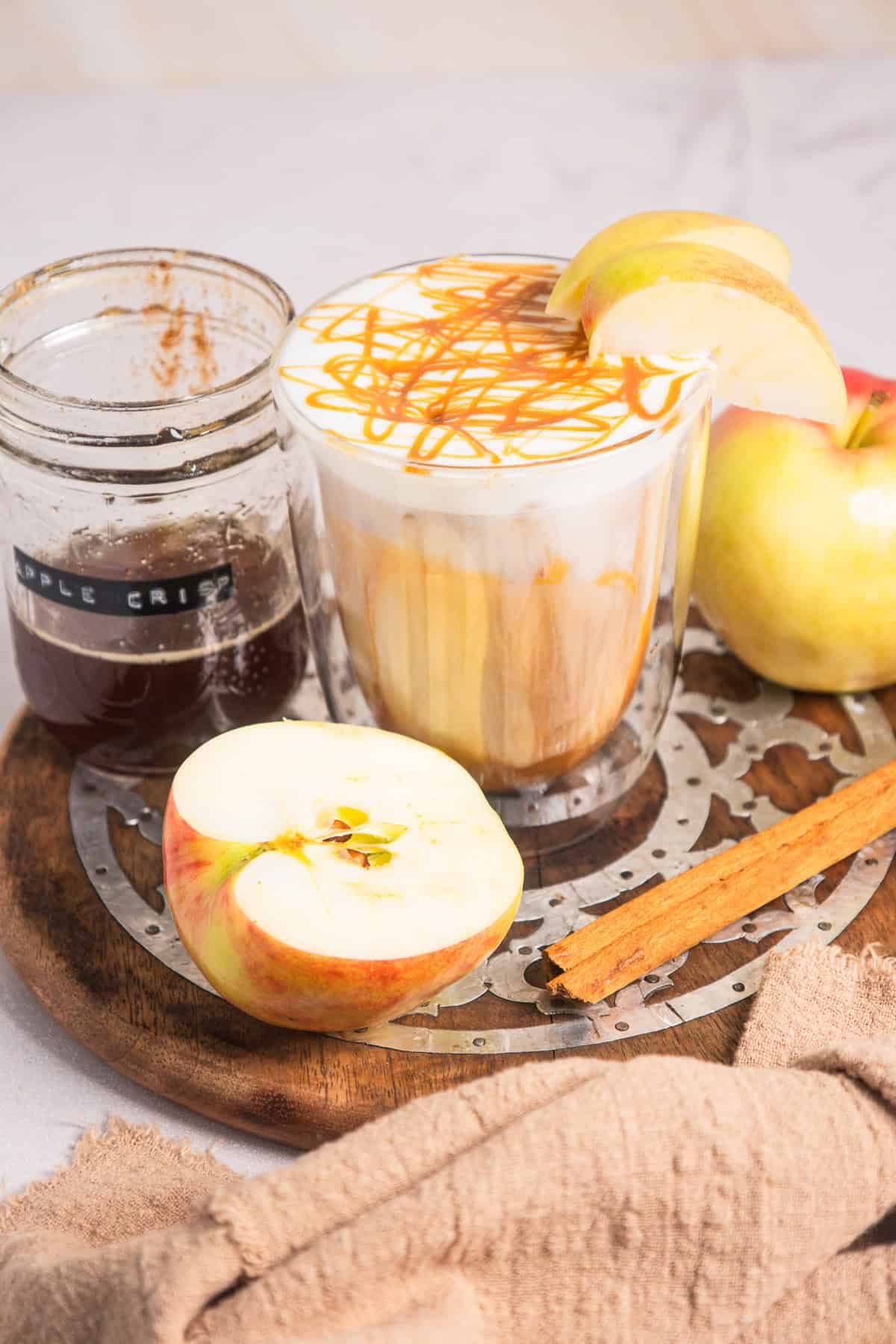 Caramel Apple Iced Coffee topped with caramel, served alongside apple slices, a cinnamon stick, and bold coffee on a wooden tray.