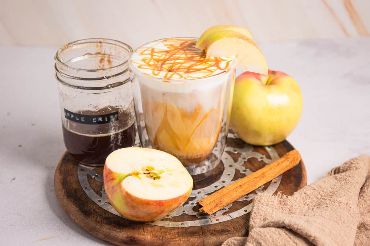 A caramel apple latte with apple slices, a jar of syrup, an apple, and a cinnamon stick on a wooden trayโperfect for fans of Caramel Apple Iced Coffee seeking cozy autumn flavors.