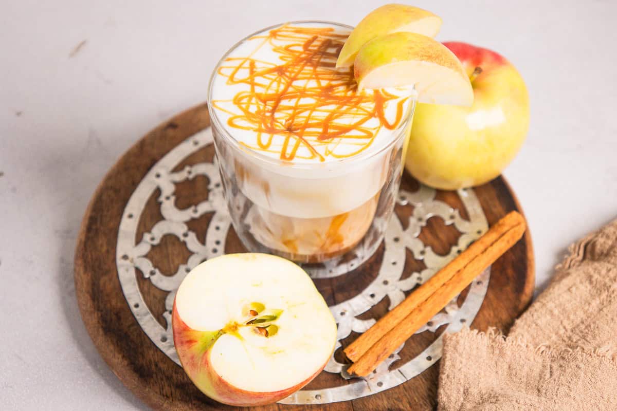 Glass of Caramel Apple Iced Coffee topped with caramel drizzle, apple slices, whole apple, and cinnamon stick nearby.