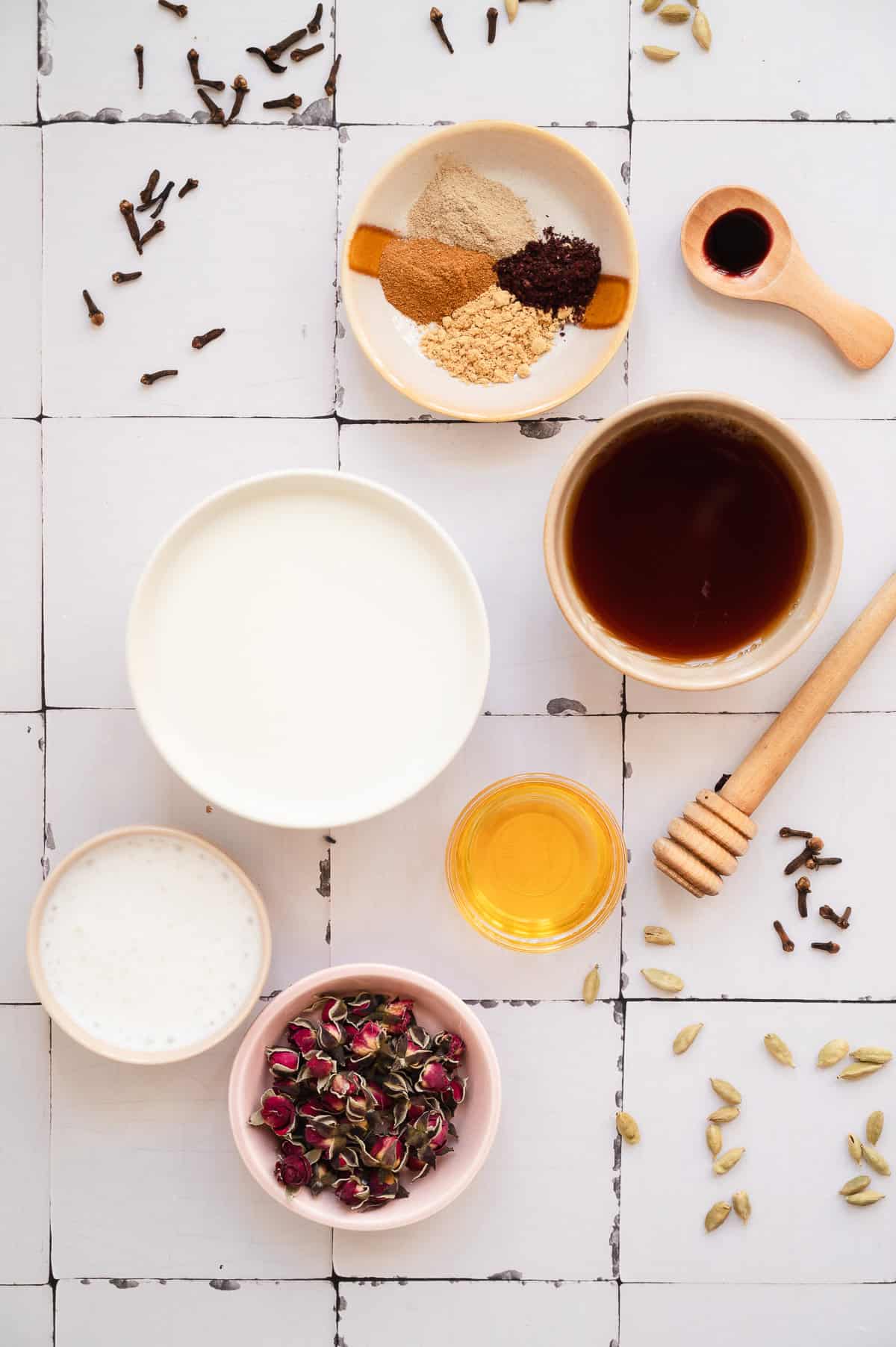 Bowls of spices, milk, tea, honey, and dried rosebuds on a white tiled surface with scattered cloves and cardamom create the perfect setting for crafting a Chai Spiced Honey Latte. Delicate dried rose petals add a fragrant finishing touch to this aromatic scene.