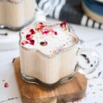 A glass mug of Chai Spiced Honey Latte with foam and dried rose petals, on a wooden coaster, beside honey and spices.