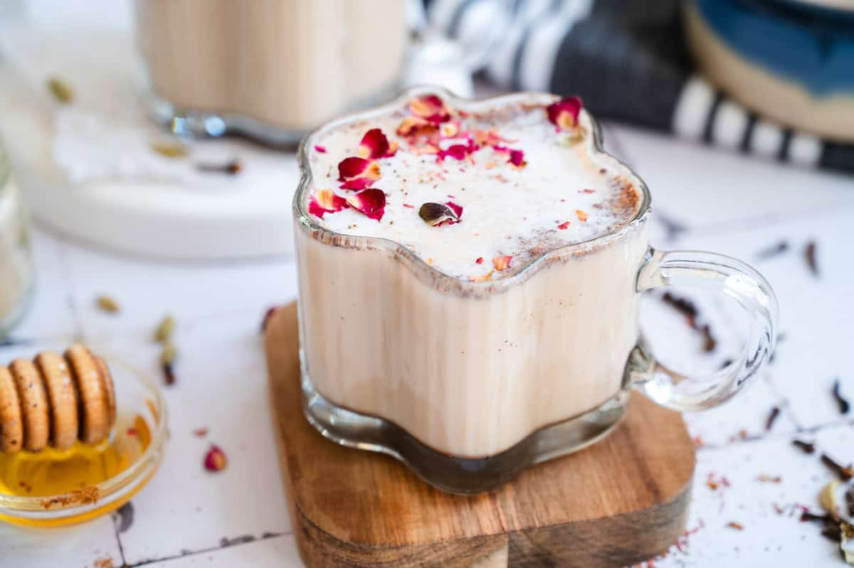 A glass of chai spiced honey latte topped with dried rose petals sits on a wooden coaster, next to honey and biscuits.