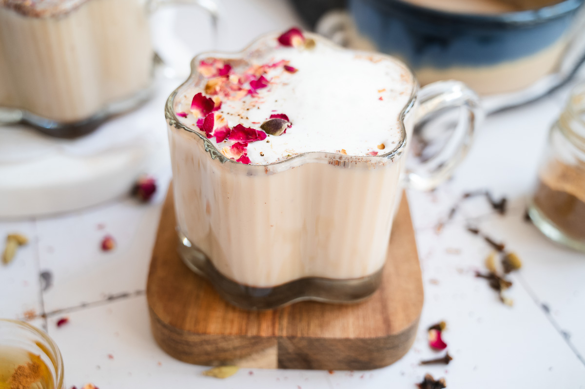 A glass mug of chai spiced honey latte with frothy milk, topped with dried rose petals, on a wooden coaster.