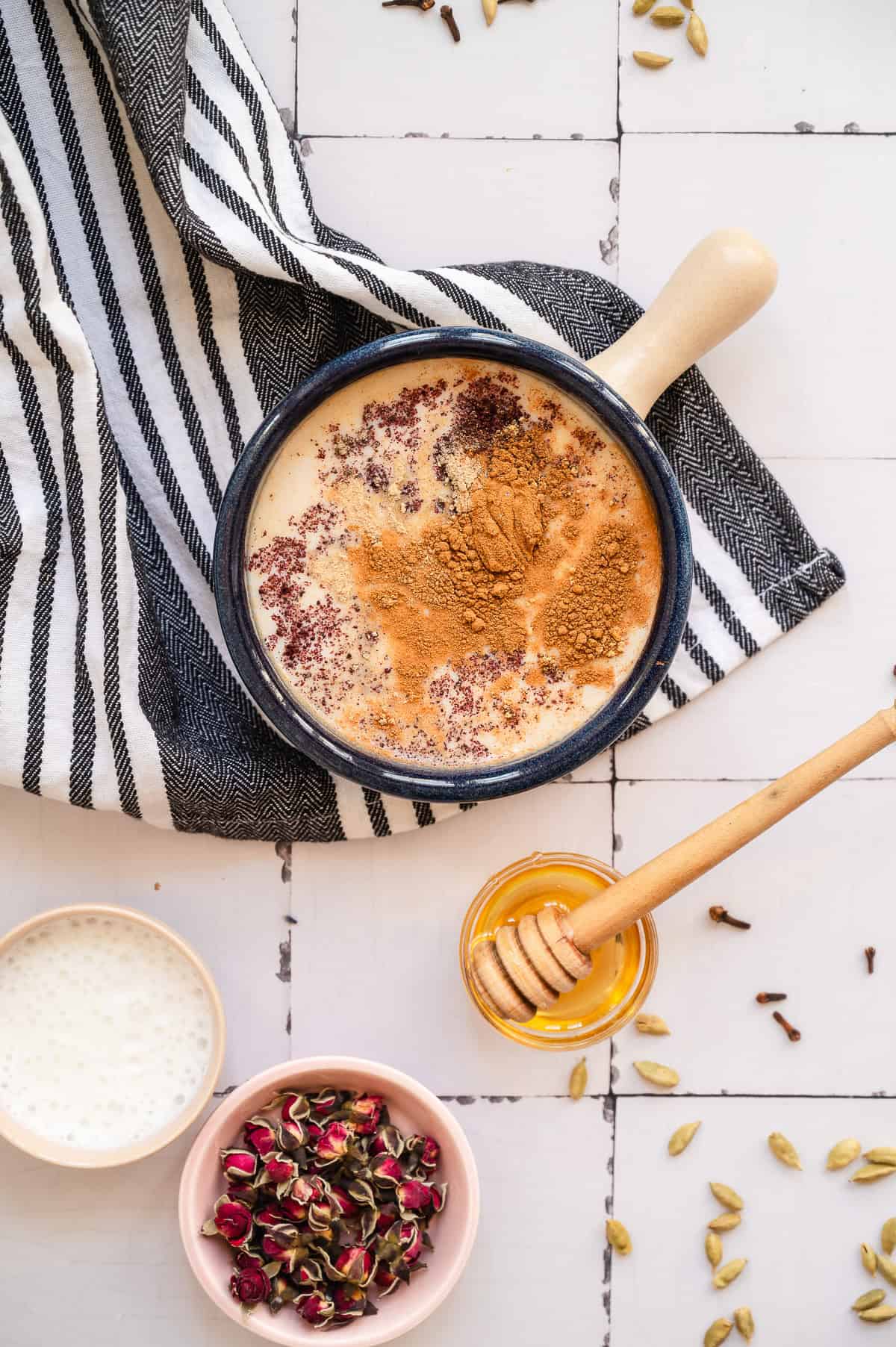 A bowl of Chai Spiced Honey Latte with cinnamon, a honey dipper, milk, dried rose petals, and spices on a striped cloth.