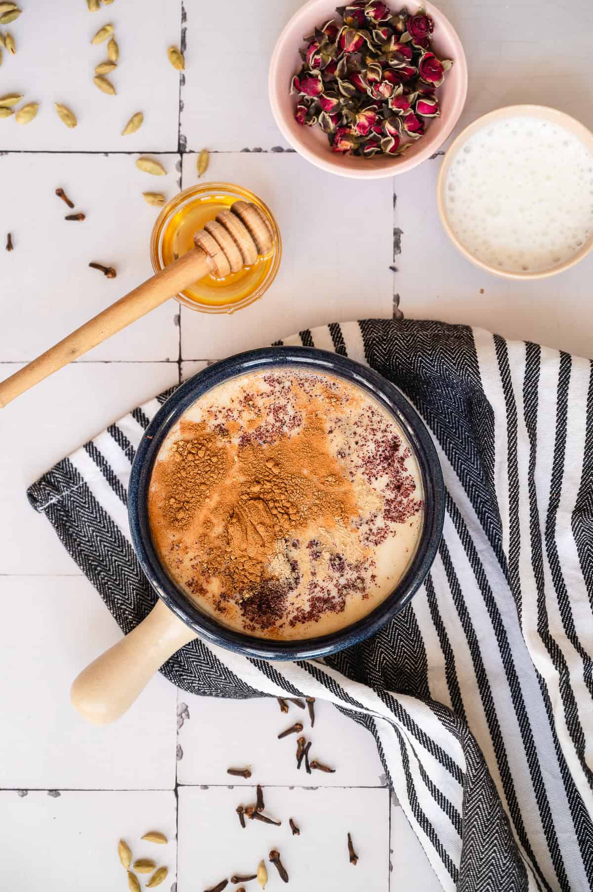 A mug of chai spiced honey latte sits on a striped cloth, with honey, dried rose petals, and milk in small bowls nearby.