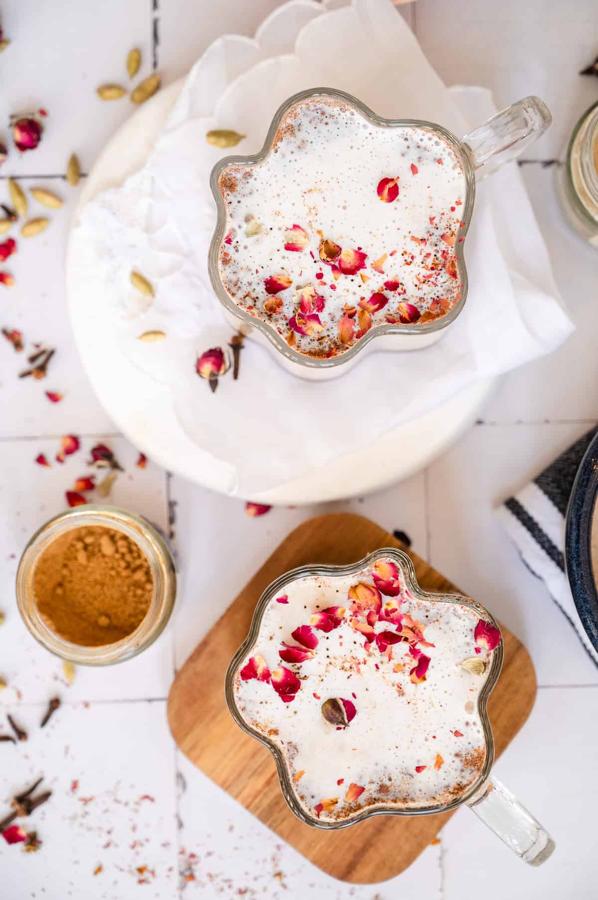 Two mugs of frothy Chai Spiced Honey Latte topped with dried rose petals and spices, surrounded by cardamom and a jar of brown sugar.