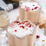 Two clear mugs of creamy Chai Spiced Honey Latte topped with frothy milk and dried rose petals, set on a light, cozy table.