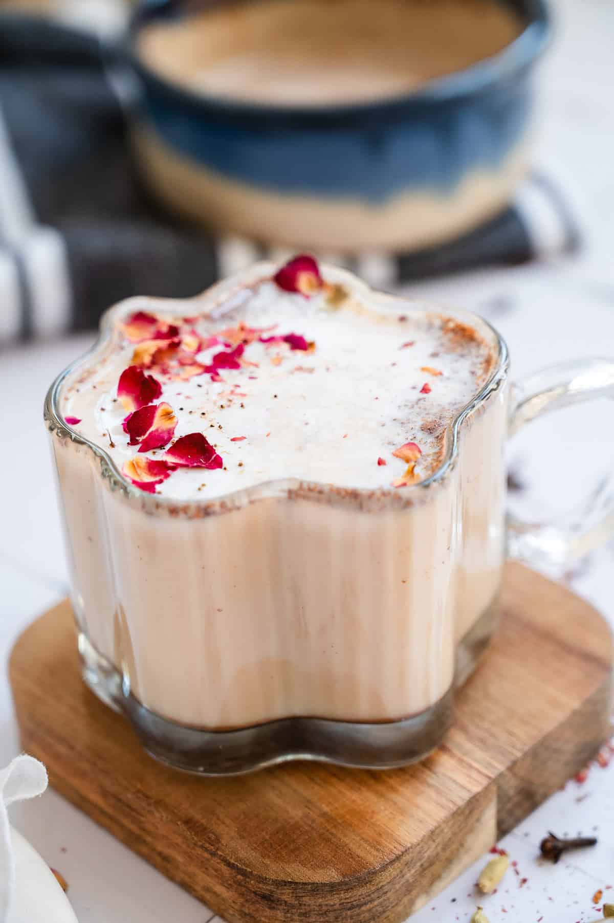 A glass mug of Chai Spiced Honey Latte topped with foam and dried rose petals sits on a wooden coaster.