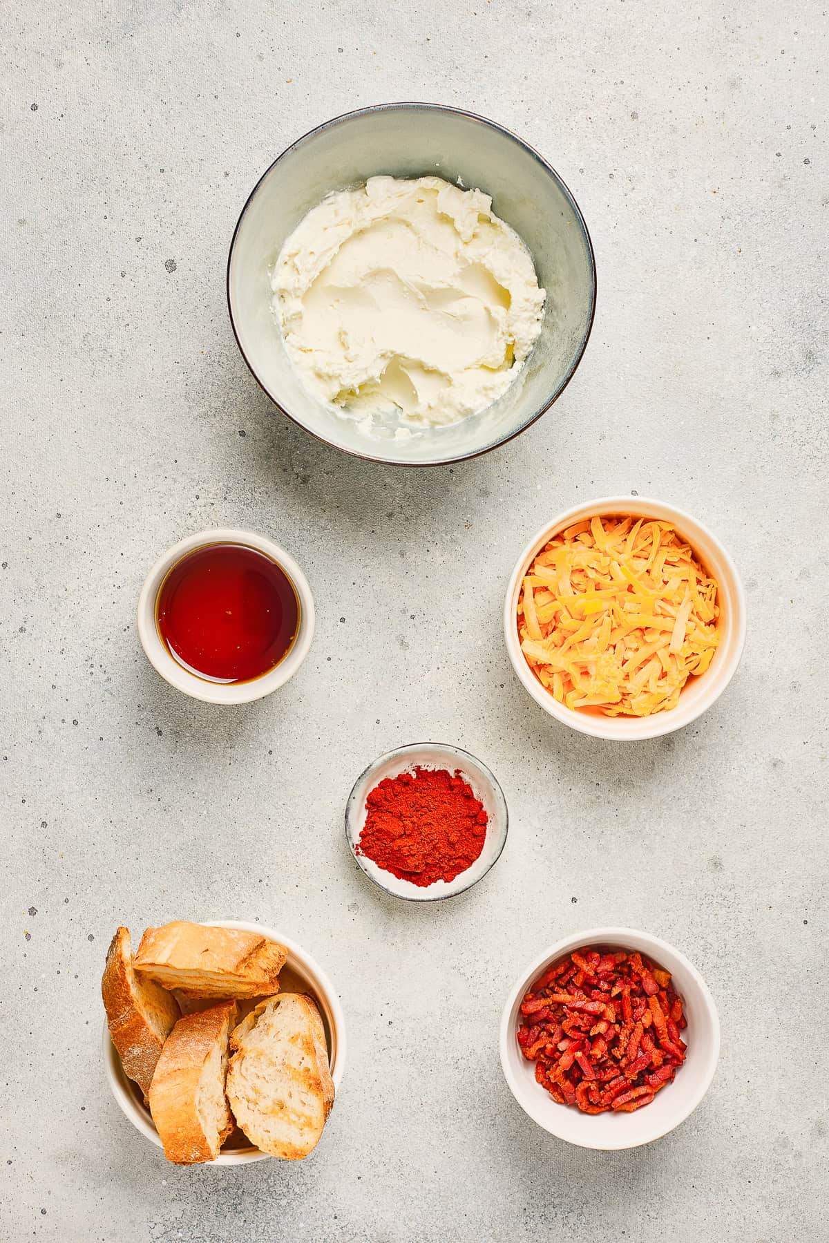 Five bowls with cream cheese, cheddar, honey, paprika, and Maple Bacon sit alongside a plate of sliced bread on a gray surface—perfect for assembling your own delicious Maple Bacon Cheese Dip.