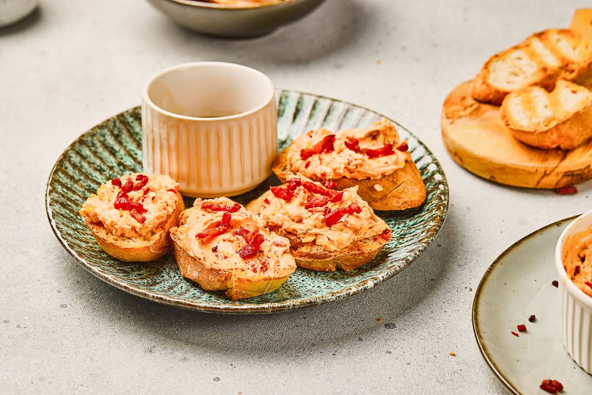 Four pieces of bread with a creamy Maple Bacon Cheese Dip and red bits on a green plate beside a white ramekin.