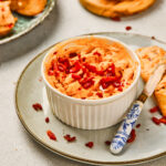 A ramekin of creamy maple bacon cheese dip topped with chopped red peppers, served with toasted bread slices on the side.