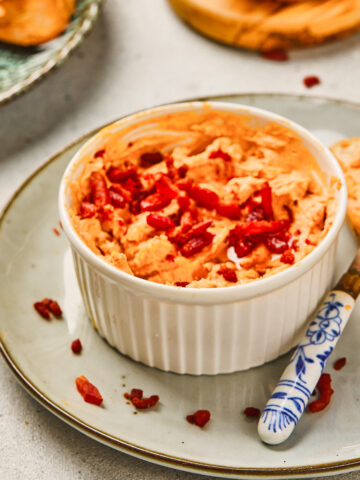 A ramekin of creamy maple bacon cheese dip topped with chopped red peppers, served with toasted bread slices on the side.