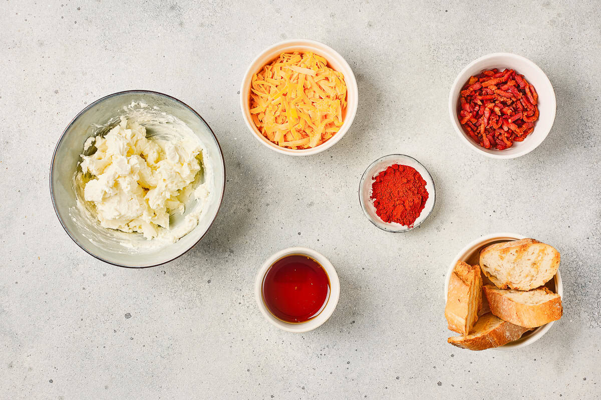 Bowls with cream cheese, shredded cheese, bacon bits, paprika, honey, and sliced bread on a light surface—perfect for creating a savory Maple Bacon Dip or Cheese Dip to share with friends.