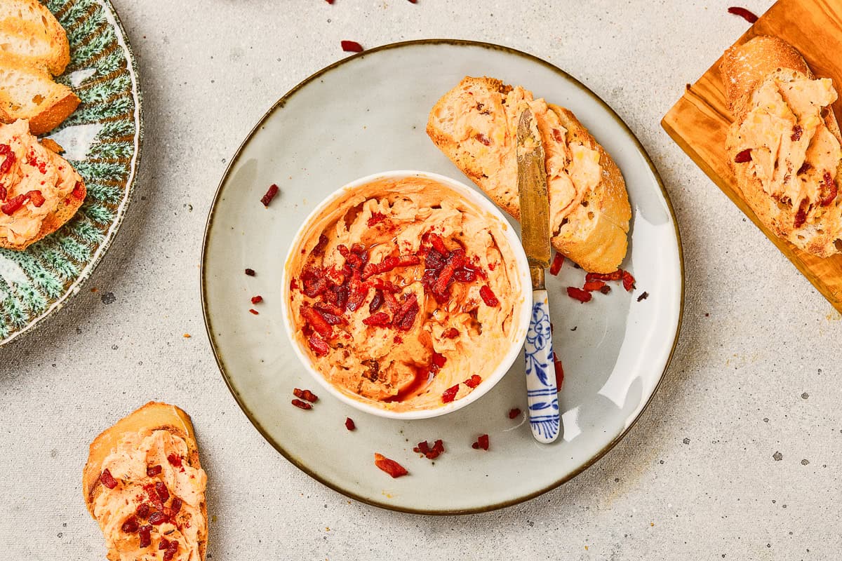 Bowl of Maple Bacon Cheese Dip topped with red bits, surrounded by bread slices on a plate and board.