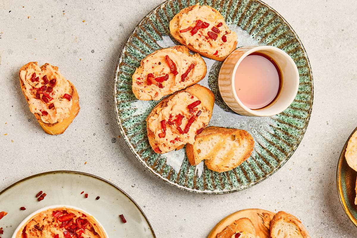 Green plate with bread slices topped with orange spread and bits of Maple Bacon Cheese, beside a small cup of pink liquid.