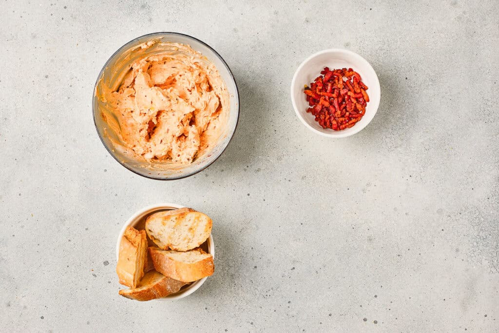 Three bowls with whipped Maple Bacon Cheese spread, chopped bacon bits, and sliced bread on a light, speckled surface.