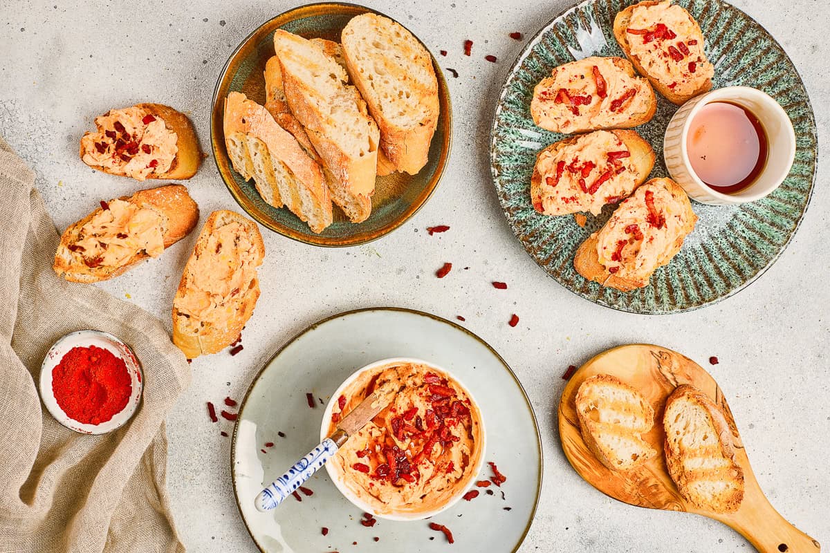 Slices of toasted bread with spread, a bowl of Maple Bacon Cheese Dip, paprika, and a cup of tea on a light surface.