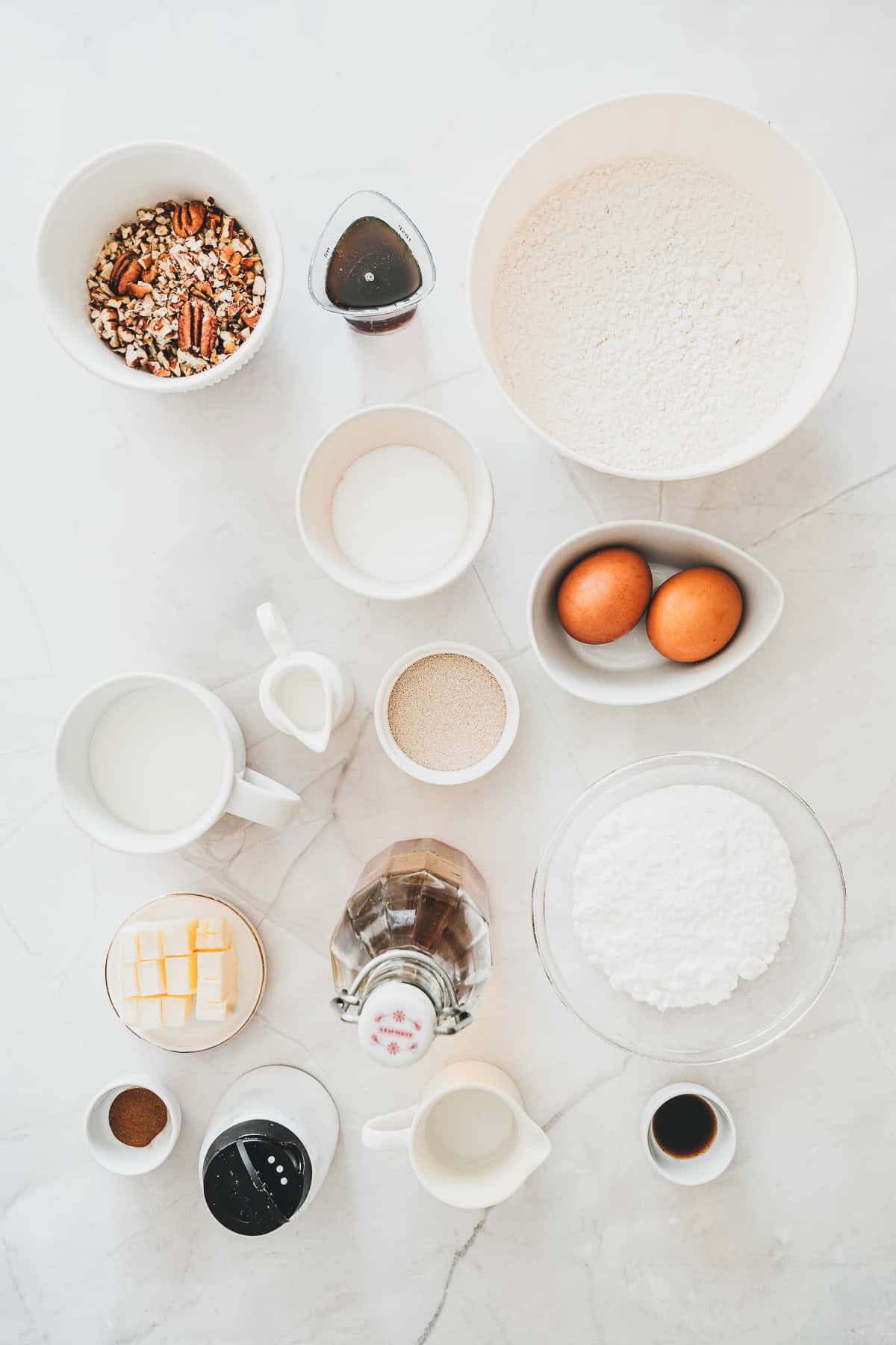 Bowls of flour, eggs, sugar, pecans, butter, milk, spices, and liquid ingredients arranged on a white surfaceโeverything you need to create delicious Maple Glazed Donuts from scratch.