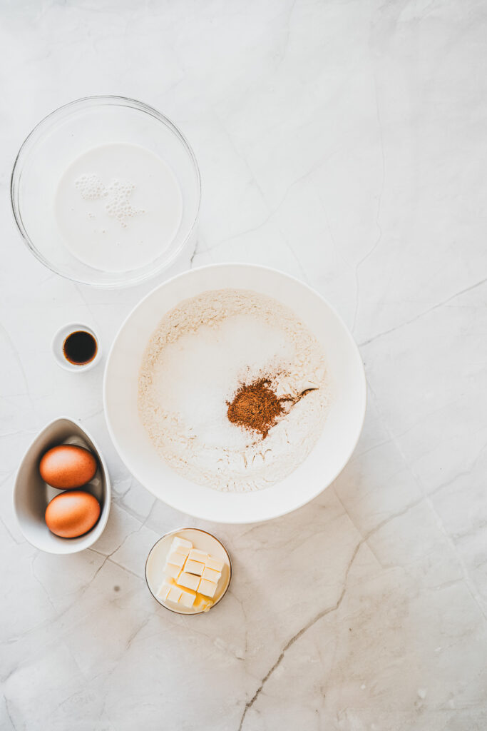 Baking ingredients on a marble surface: flour, eggs, butter, vanilla, cinnamon, and milk in separate bowlsโeverything you need to make delicious Maple Glazed Donuts from scratch.