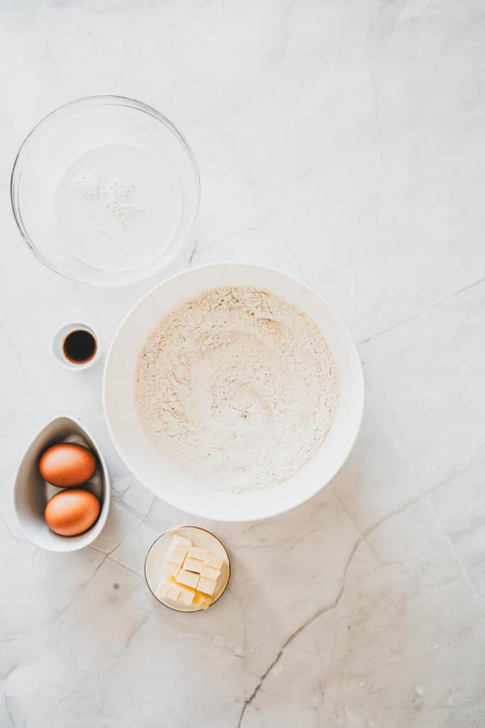 Baking ingredients on a white surface: flour, eggs, butter cubes, vanilla, and milk in bowlsโperfect for creating delicious Maple Glazed Donuts.