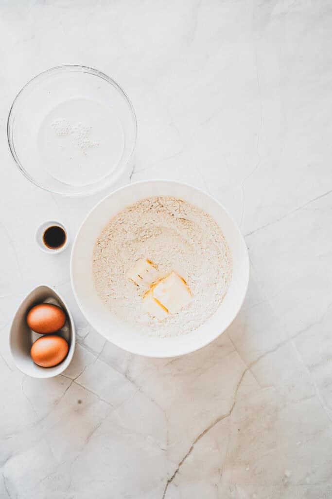 On a marble counter, a bowl of flour with butter sits beside two eggs, vanilla, and a bowl of waterโsimple ingredients ready to be transformed into delicious Maple Glazed Donuts.