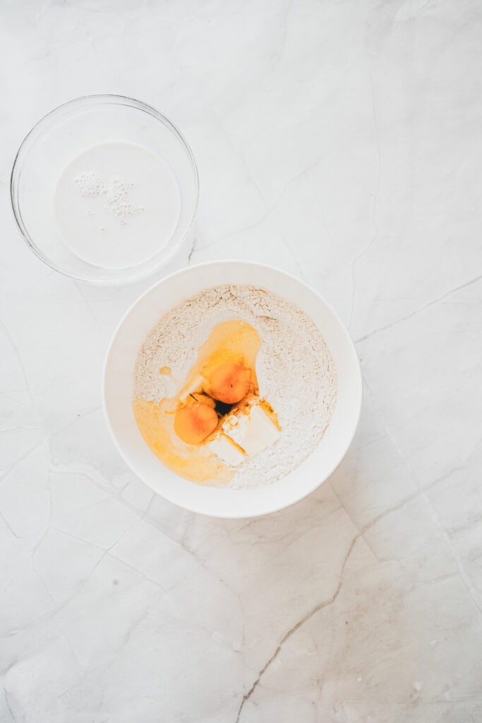 A bowl with flour, eggs, and butter next to a bowl of yeast on a white marble surfaceโperfect essentials for making homemade Maple Glazed Donuts.