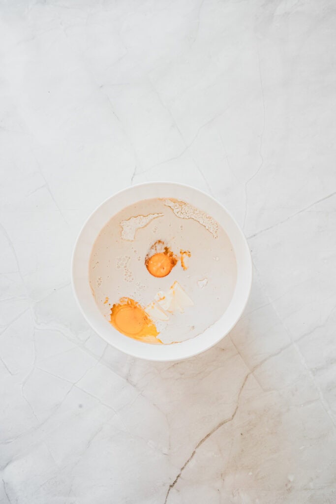 A white bowl with eggs, flour, and milk on a light marble surface, viewed from aboveโperfect ingredients for making delicious Maple Glazed Donuts at home.