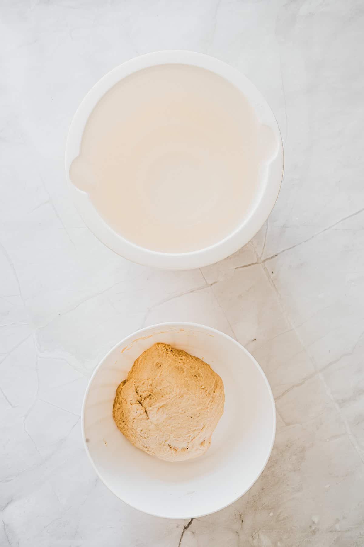 Two white bowls rest on a marble surface, one empty and the other cradling a ball of doughโperfect for making fresh maple glazed donuts.
