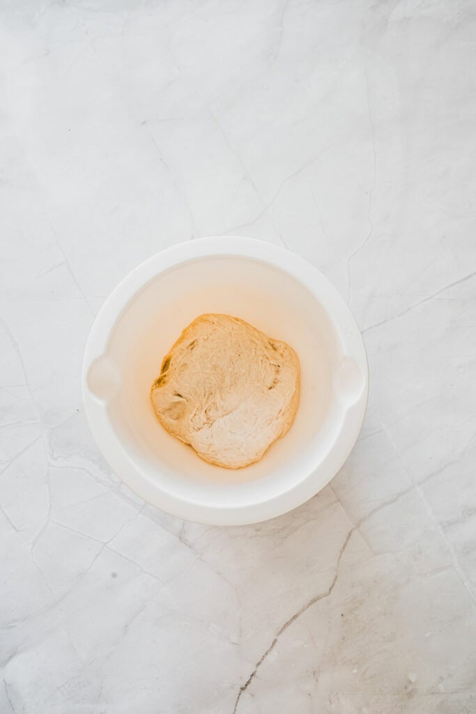 A ball of dough, soon to become delicious Maple Glazed Donuts, rests in a white bowl on a light marble surface.